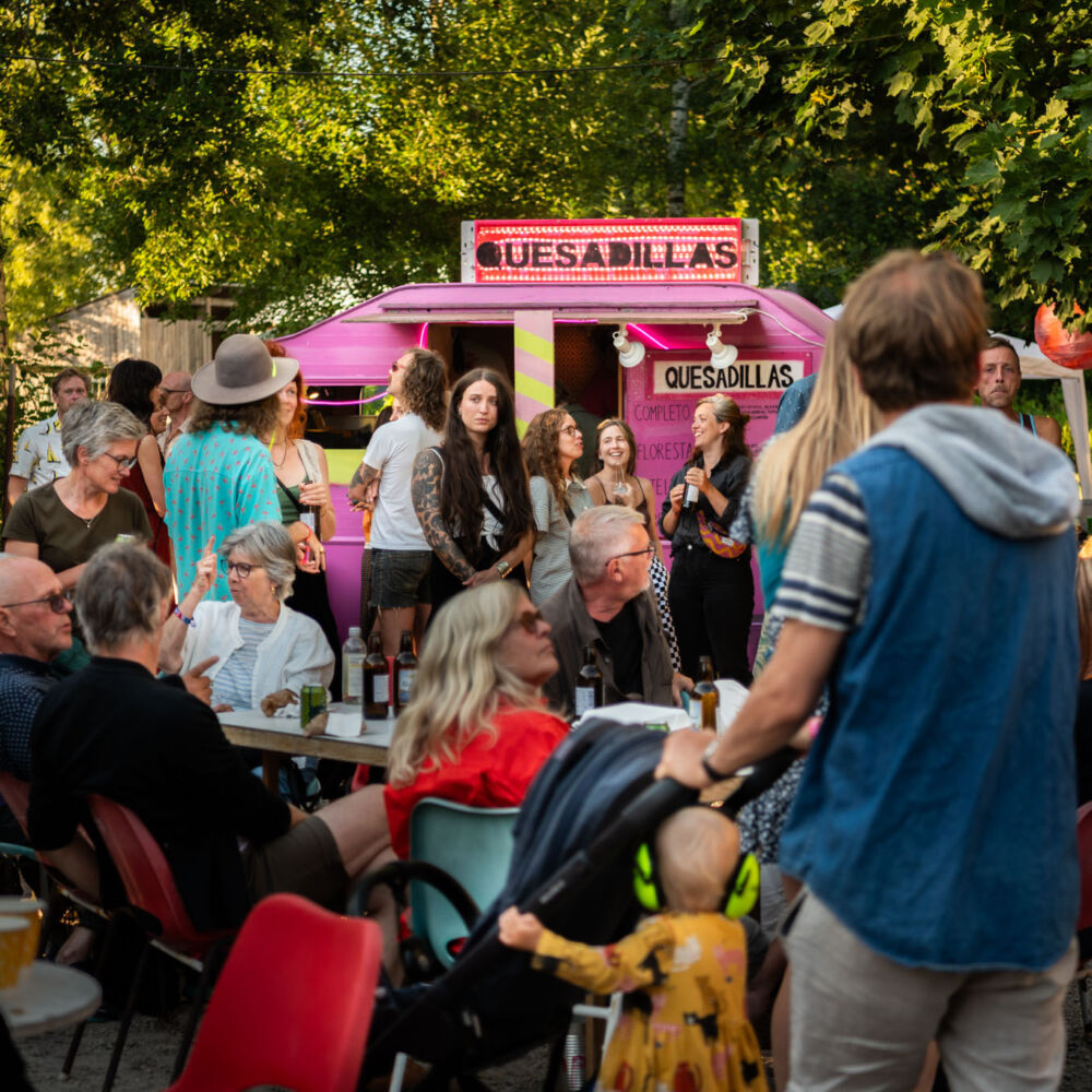 Människor samlas vid en rosa foodtruck under en sommarkväll med musik och servering.